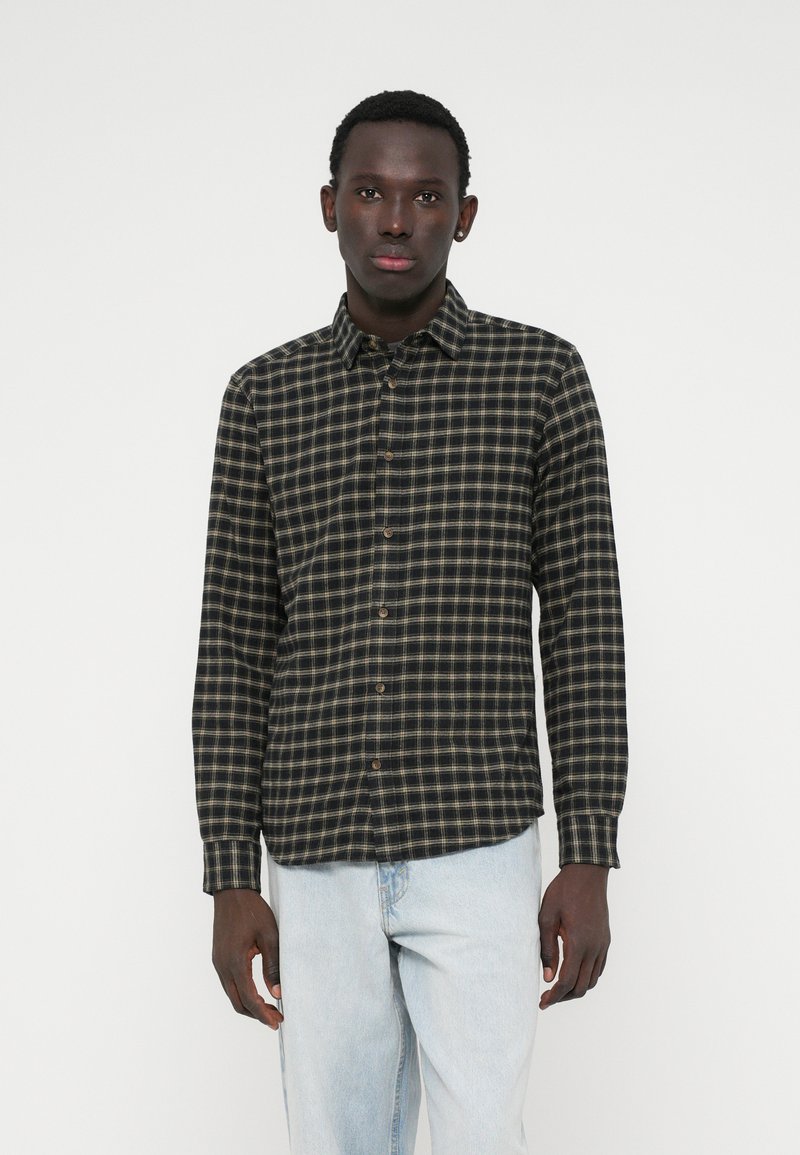 Νεαρός άνδρας με σκούρο δέρμα, φορώντας ένα μαύρο και μπεζ καρό πουκάμισο με μακριά μανίκια και ανοιχτό μπλε τζιν, στέκεται μπροστά σε λευκό φόντο.