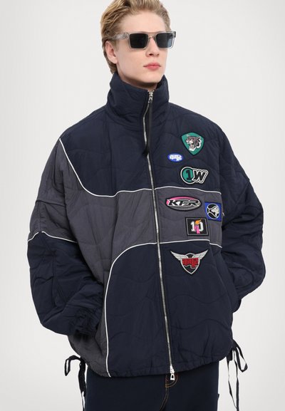 Padded navy jacket with contrasting gray sections, featuring various embroidered patches and a high collar. Drawstring hem and zip closure.