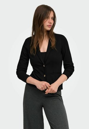 Femme aux longs cheveux bruns portant un blazer noir et un pantalon gris, debout les mains jointes, regardant sur le côté sur un fond uni.