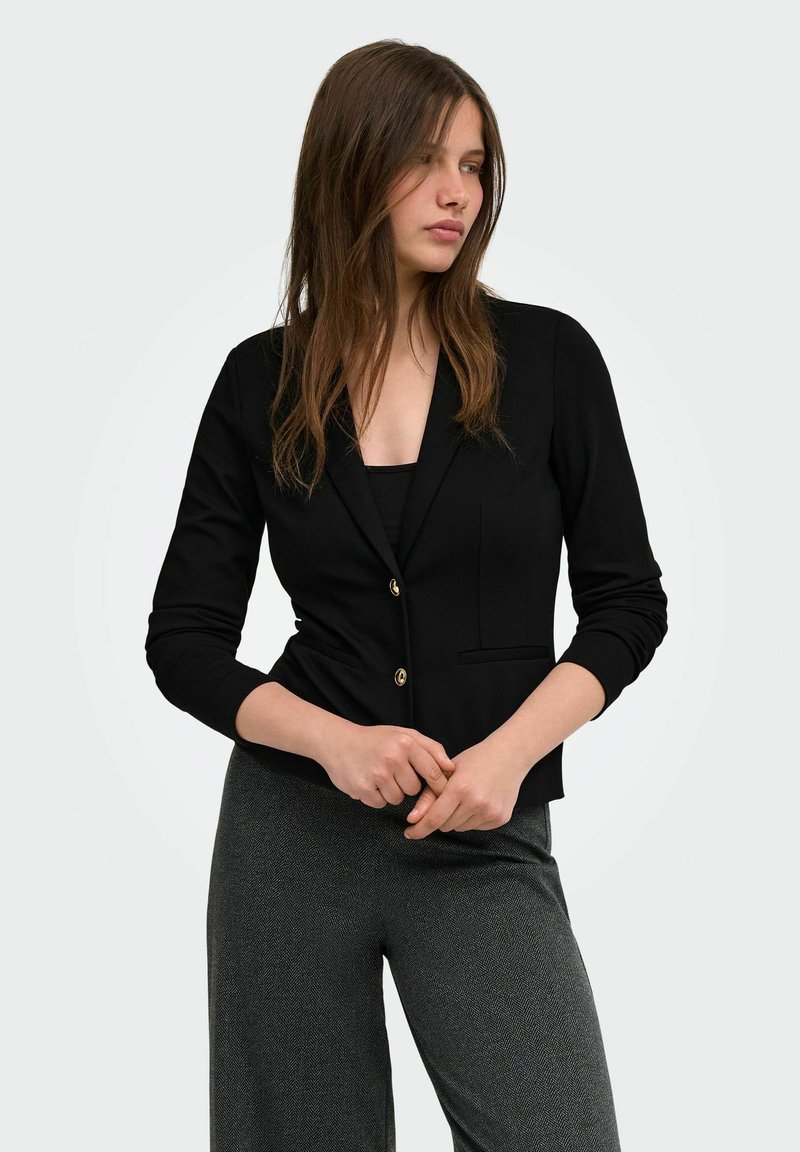Femme aux longs cheveux bruns portant un blazer noir et un pantalon gris, debout les mains jointes, regardant sur le côté sur un fond uni.