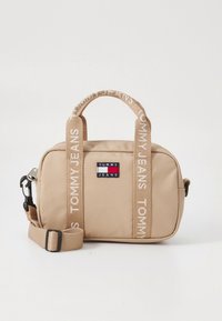 Sac bandoulière beige Tommy Jeans avec double poignée et bandoulière réglable, arborant le logo Tommy Jeans à l'avant.