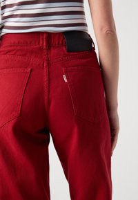 Personne portant un pantalon en jean rouge et une chemise rayée blanche, marron et violette, vue de la taille jusqu'au milieu de la cuisse de dos.