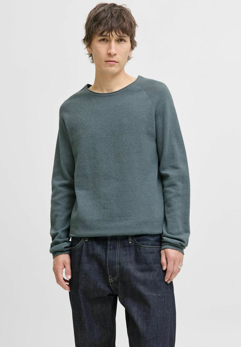 Tricou verde texturat cu un decolteu larg și mâneci lungi, purtat cu blugi negri din denim. Design simplu fără modele sau accente vizibile.