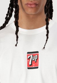 Hvid t-shirt med korte ærmer, som har et 7-Up logo i rød og sort på forsiden, med en glat stoftekstur.
