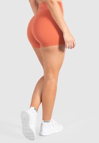 Pantaloni sportivi arancioni con un design aderente, texture liscia e vita media, abbinati a sneakers bianche con suola texturizzata.
