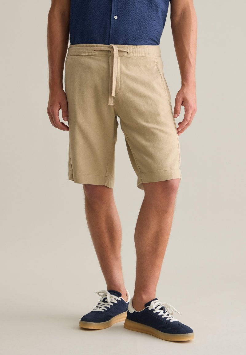 Shorts beiges avec une taille à cordon et des poches latérales. Associés à des baskets en daim bleu marine avec des semelles en caoutchouc blanc et gomme.