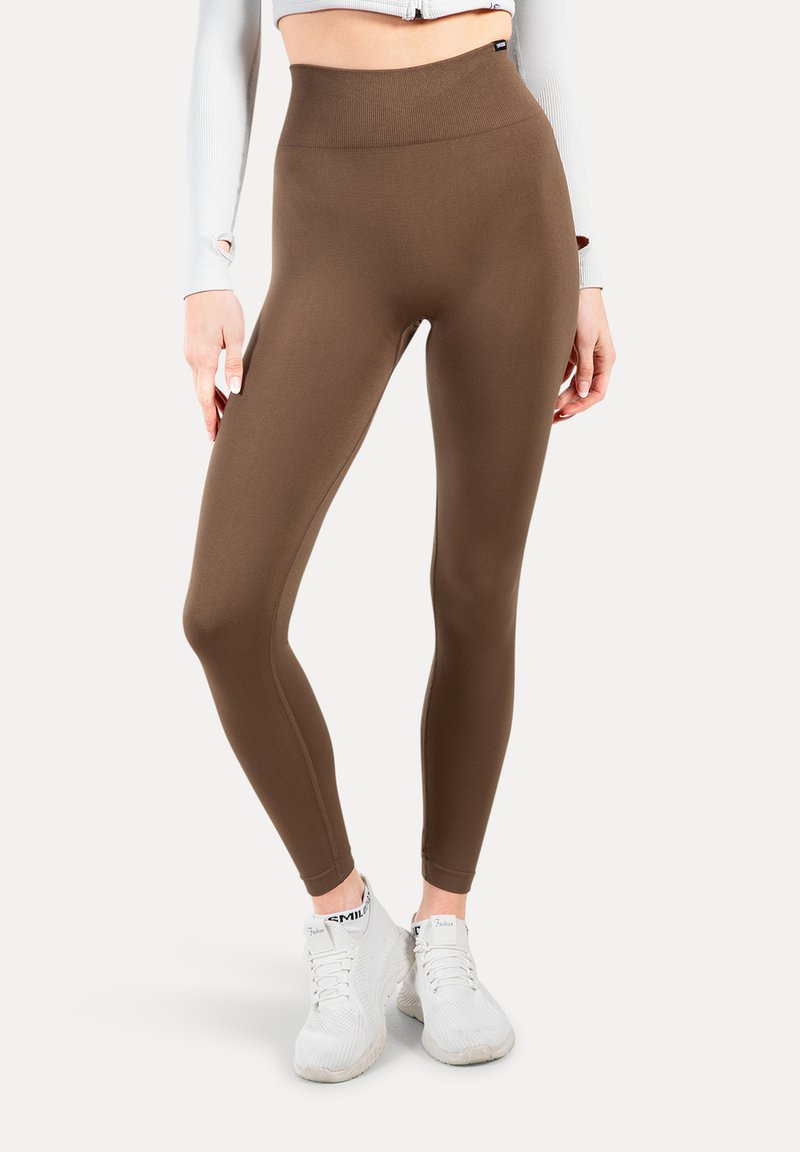 Bruine high-waisted leggings van rekbare stof. Soepele textuur, figuurvormend ontwerp en enkel lengte. Gecombineerd met witte sportschoenen.