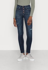 Höga, mörkblå denimjeans med slitna detaljer och fem-knappsstängning, parat med grå sneakers. Slät textur, åtsittande form.
