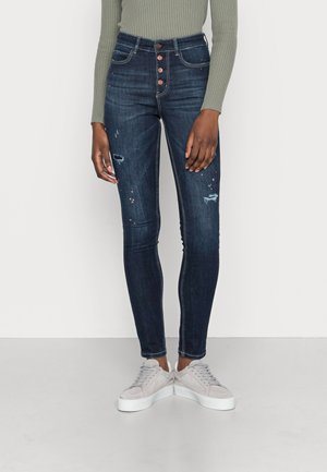 High-waisted dark blue denim jeans with distressed details and five-button closure, paired with gray sneakers. Smooth texture, fitted shape.