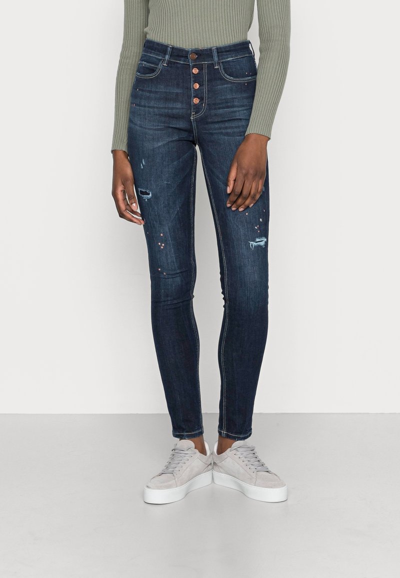 Höga, mörkblå denimjeans med slitna detaljer och fem-knappsstängning, parat med grå sneakers. Slät textur, åtsittande form.