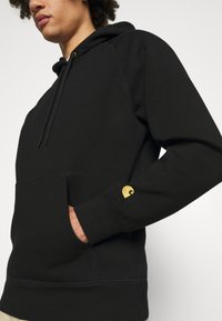 Musta huppari, jossa on etutasku, ribbireunat ja kultainen logo yksityiskohtana hihassa. Pehmeä kangas, rento istuvuus.