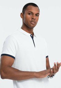 Polo shirt bianco con bordo blu navy sul colletto e sulle maniche. Il tessuto sembra avere una texture. L'individuo sta sistemando le mani.