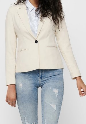 Femme portant un blazer crème à un seul bouton sur une chemise rayée bleue et un jean bleu clair déchiré, debout avec les mains détendues le long du corps.