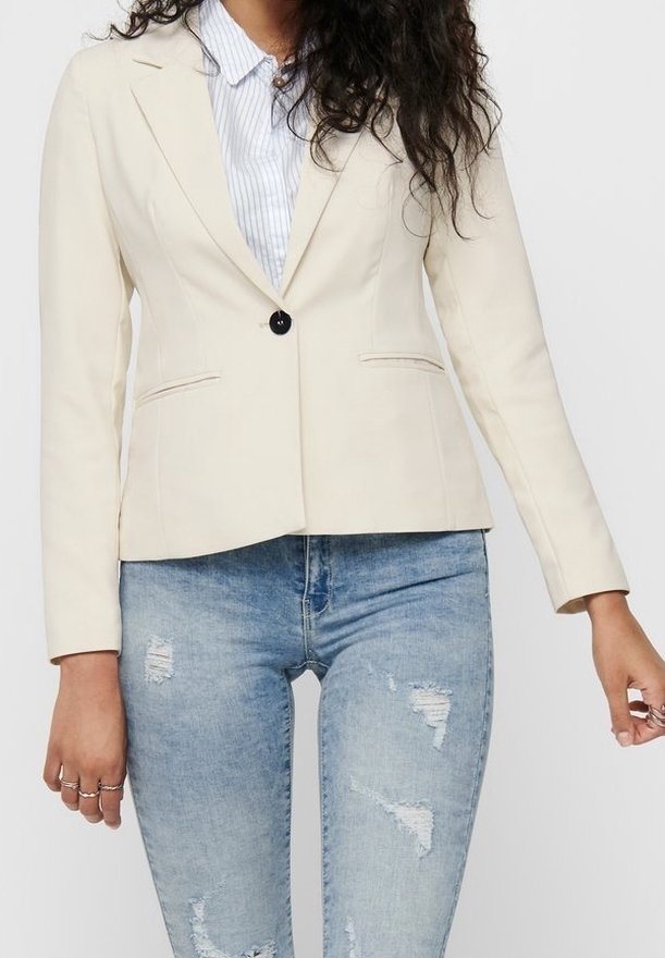 Femme portant un blazer crème à un seul bouton sur une chemise rayée bleue et un jean bleu clair déchiré, debout avec les mains détendues le long du corps.