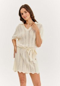 Sukienka dzianinowa w jasnym kremie z krótkimi rękawami, dekoltem w kształcie litery V i paskiem zawiązanym wstążką. Posiada falbankowy dół i otwarty wzór.