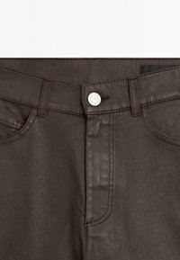 Pantalon marron foncé avec fermeture à boutons sur le devant, passants de ceinture et poches latérales, montrant des coutures détaillées autour de la taille.