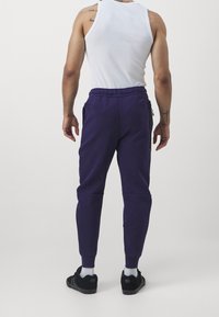 Lila sweatpants med dragsko i midjan, sidofickor och smalnande anklar, tillsammans med en vit linne och svarta sneakers.