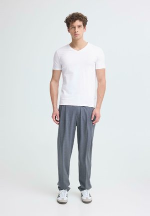 Giovane uomo con capelli ricci che indossa una maglietta bianca a V semplice, pantaloni grigi plissettati e sneakers bianche, in piedi davanti a uno sfondo bianco.