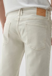 Helle beige Denim-Hose mit glatter Textur, ausgestattet mit Gesäßtaschen und einem braunen Leder-Logo-Patch am Bund.
