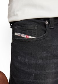 Jeans de mezclilla negra con un acabado texturizado, una etiqueta contrastante con acentos rojos visible en el bolsillo delantero y un cierre de botón de metal.