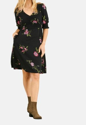 Kvinde iført en sort, blomsterprintet knælængde kjole med pufærmer og brune ankelstøvler, stående med den ene hånd i lommen.