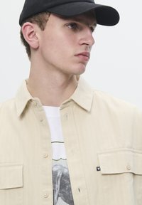 Beige button-up shirt with a pocket, worn over a white graphic T-shirt featuring elephant print. Black cap adds contrast to the outfit.