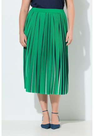 Kvinde iført en grøn plisseret midi-nederdel med vertikale marineblå og hvide striber, kombineret med marineblå ankelremspointede sko.