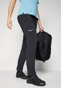 Calças de exercício pretas com bolsos cargo, apresentando um logótipo branco, combinadas com uma camisa azul e uma mochila preta com detalhes texturizados.