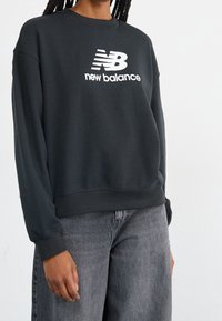 Felpa nera con collo a girocollo, caratterizzata dal logo bianco "New Balance". Tessuto morbido, maniche lunghe e orlo a coste. Indossata con jeans grigi.