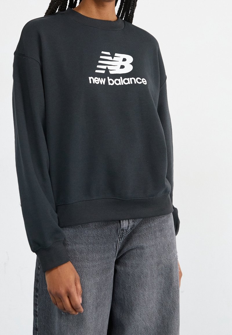 Felpa nera con collo a girocollo, caratterizzata dal logo bianco "New Balance". Tessuto morbido, maniche lunghe e orlo a coste. Indossata con jeans grigi.