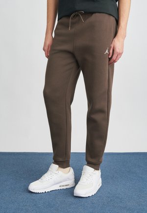 Brown sweatpants with a drawstring waist, featuring a white logo on the side, paired with white sneakers on a blue carpet background.