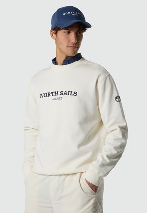 Jeune homme portant un sweat-shirt crème avec le texte « North Sails Archive » et un pantalon assorti, une casquette bleue, les mains dans les poches, regardant vers la gauche.