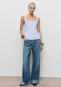 Weißes Top mit quadratischem Ausschnitt, hellblaue weite Jeans mit umgeschlagenen Säumen und schwarze High Heels. Akzentuiert mit klobigen Armreifen an einem Arm.