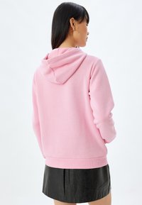 Sudadera con capucha rosa, fabricada con un tejido suave, que presenta un corte relajado, puños acanalados y un bolsillo tipo canguro. Lleva puesta sobre una falda negra.