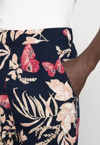 Blommönstrade byxor med en mörk marinblå bakgrund, stora rosa fjärilar och beige blad. Sidoficka synlig med textilstruktur.