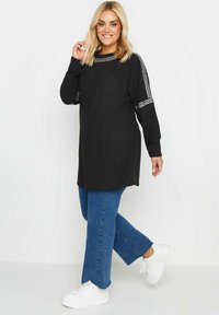 Svart tunika topp i strukturerad tyg, med rund halsringning och långa ärmar. I kombination med blå raka jeans och vita sneakers.