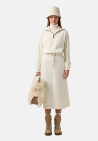 Cremfarbener, gesteppten Oberteil mit Reißverschluss, passender Midi-Rock, beige Stiefel mit Fellbesatz und eine gesteppten Bucket Hat. Hält eine mit Fleece gefütterte Tasche.