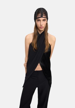 Kobieta o długich włosach, ubrana w czarny, bezrękawnikowy, zapinany top, czarne spodnie oraz czarną opaskę z koralików na głowie, na jasnym, jednolitym tle.