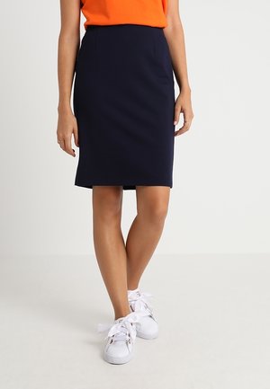 Woman wearing a navy knee-length skirt, orange top, and white sneakers standing with legs crossed on a neutral background.