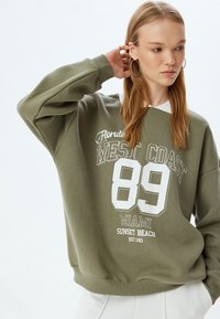 Sudadera verde oliva con texto impreso en blanco, presenta un escote redondeado, puños acanalados y un corte holgado. Material de mezcla de algodón, textura suave.