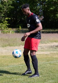Camiseta de fútbol negra de manga corta con logo, detalles rojos, combinada con pantalones cortos rojos, calcetas negras y botas negras, en un campo de hierba.