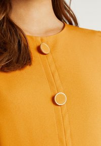 Tejido naranja con una textura suave que presenta dos botones metálicos redondeados. Los botones tienen un patrón sutil y están situados en la parte delantera.