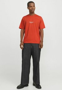 Rotes Kurzarm-T-Shirt mit dem Aufdruck "Originals Studio" auf der Brust, kombiniert mit locker sitzenden dunklen Jeanshosen und schwarzen Schuhen.