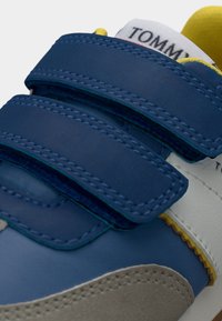 Μπλε και γκρι αθλητικά παπούτσια με λουράκια βέλκρο, λείες και υφασμένες επιφάνειες, με κίτρινες λεπτομέρειες και αντίθετες ραφές.