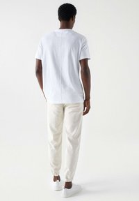 Camiseta blanca de algodón con corte recto, detalle en la costura trasera, combinada con pantalones joggers tapered en un beige claro y zapatillas blancas. Diseño minimalista.