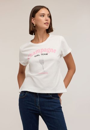 Femme aux cheveux courts et bruns, portant des boucles d'oreilles créoles dorées, un t-shirt blanc avec le texte "champagne girl team", et un jean bleu, posant les mains dans les poches.