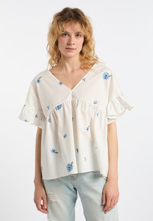 Junge Frau mit lockigem blondem Haar, die eine weiße Bluse mit blauen Blumenmustern und hellblaue zerrissene Jeans trägt, steht vor einem weißen Hintergrund.