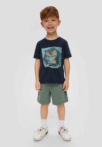 Marinefarbener Baumwoll-T-Shirt mit einem blauen und orangefarbenen Grafikdruck, kombiniert mit grünen Shorts. Weiße Sneakers mit schwarzen und roten Akzenten. Kurze Ärmel.