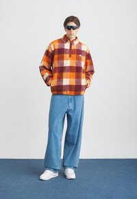 Lodden, rutete fleece-pullover i oransje, hvitt og burgunder med halv-zip krage, parret med løse lyseblå jeans og hvite sneakers.