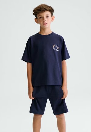 Ragazzo in piedi con indosso una T-shirt blu scuro e pantaloncini, la T-shirt con una piccola grafica di calcio e testo sul petto, sfondo chiaro e semplice.